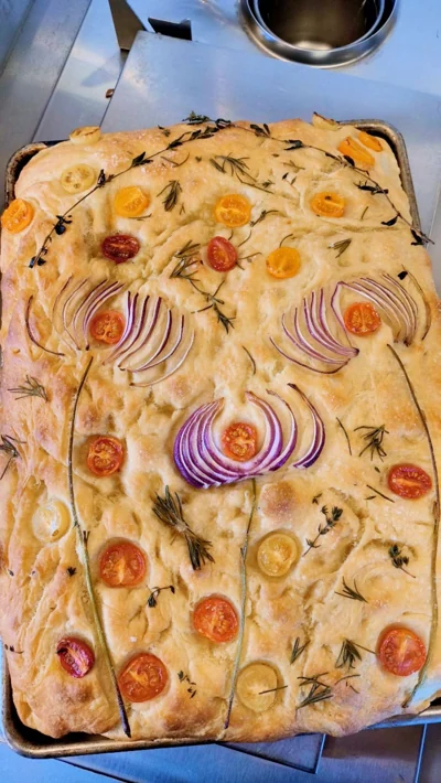 Focaccia decorated with tomatoes, red onion, rosemary, and thyme.